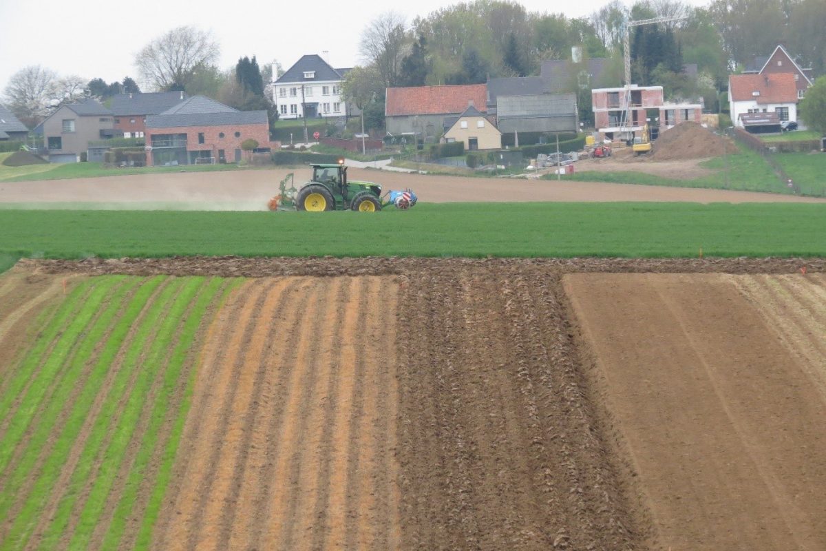 Op de voorgrond licht afhellende percelen ingezaaid met de helling mee, op de achtergrond rijdt een tractor voorbij