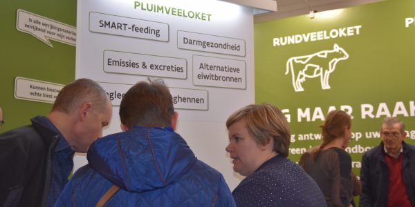Three people talking at a trade show in front of a poster saying "pluimveeloket"