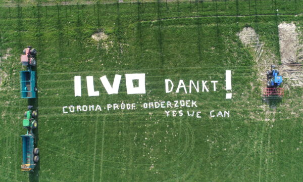 Dronefoto van een akker met gras met de tekst: ILVO dankt! Corona-proof onderzoek yes we can. De tekst is geflankeerd door 3 tractoren met aanhangwagens.