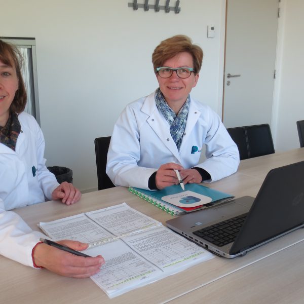 Two smiling women in lab coats seated at a table with a computer and papers