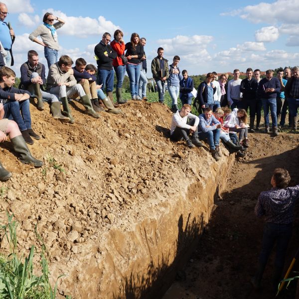 Demonstratie van: Het herkennen van een gezonde bodem, hoe ga je te werk? Een man staat in een gegraven put met een dertigtal toeschouwers.