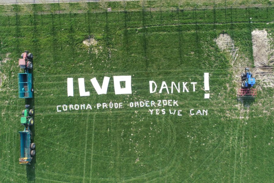 Dronefoto van een akker met gras met de tekst: ILVO dankt! Corona-proof onderzoek yes we can. De tekst is geflankeerd door 3 tractoren met aanhangwagens.